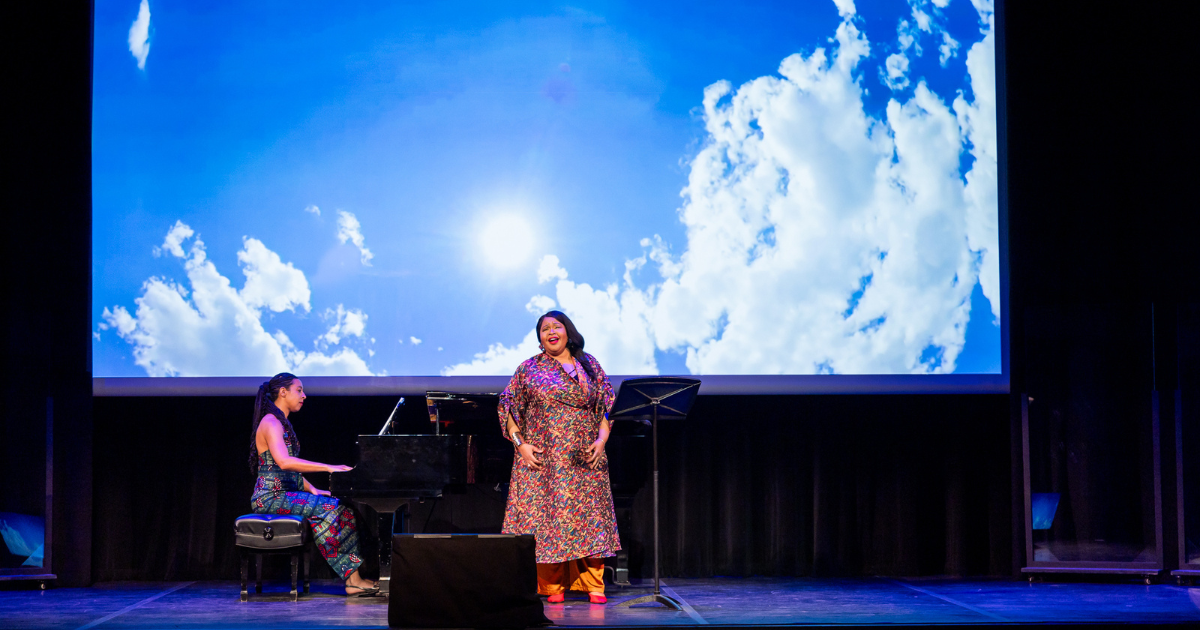 2024-03-27 Karen Slack Closes Out Intensive Residency with Standing Ovation A performer sings on stage with a pianist and clouds in the background