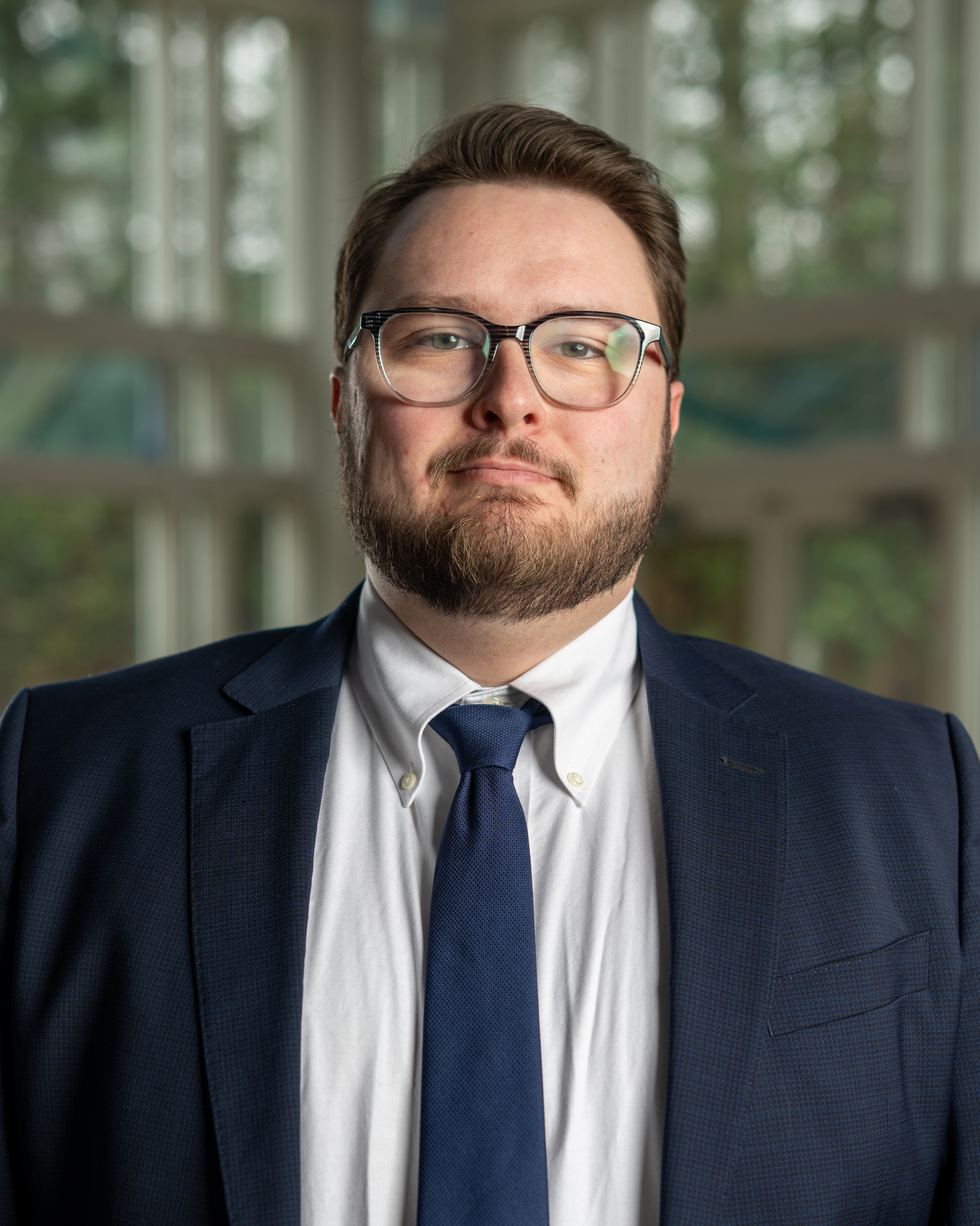 Charles has brown hair and is wearing a blue suite jacket, white undershirt and blue tie with glasses in front of a multi-color background.